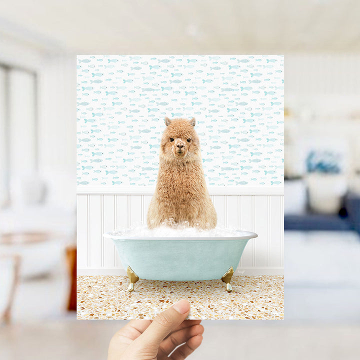 A hand holds up a framed image of a brown alpaca sitting in a blue bathtub filled with bubbles, surrounded by a pattern of fish.