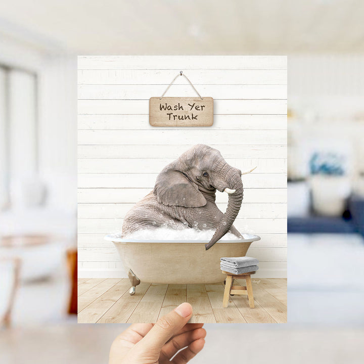 A hand holds up a framed image depicting a gray elephant sitting in a bathtub filled with water, with a sign above that reads "Wash Your Trunk".