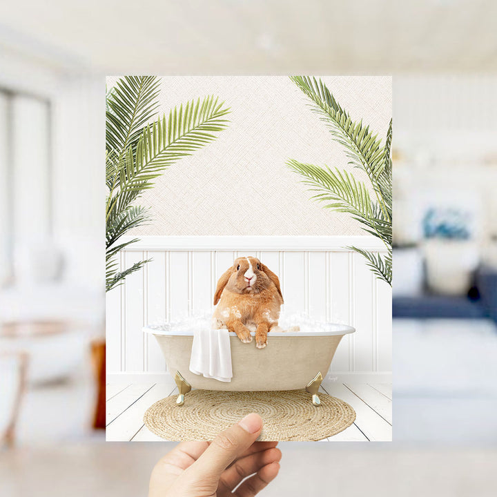 A hand holds up a framed photograph of a brown rabbit sitting in a bathtub filled with water, surrounded by green palm leaves.