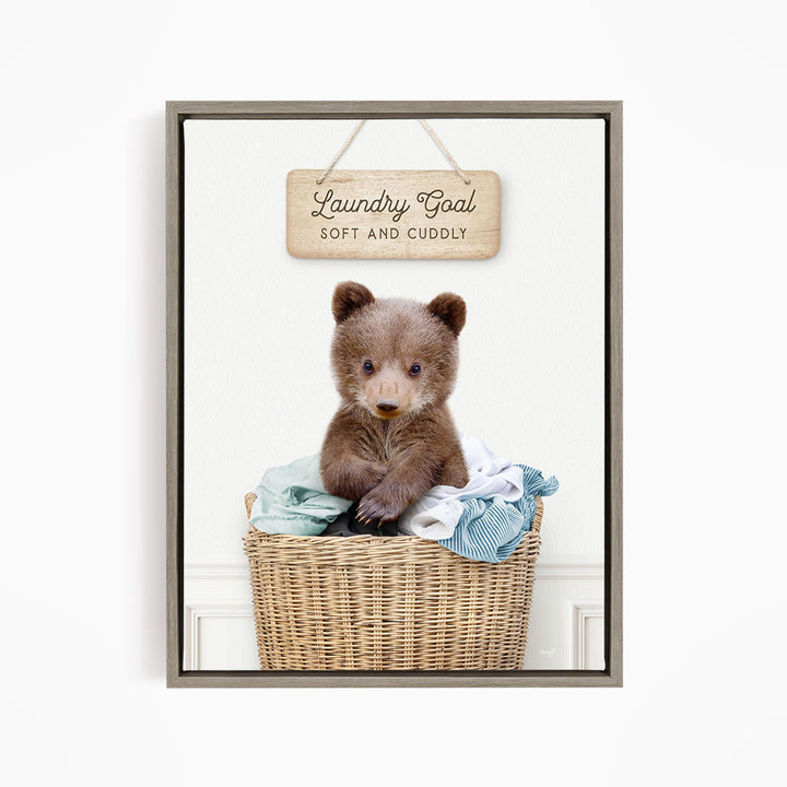 A cute brown bear sitting in a wicker laundry basket, with a wooden sign above that says "Laundry Goal Soft and Cuddly".