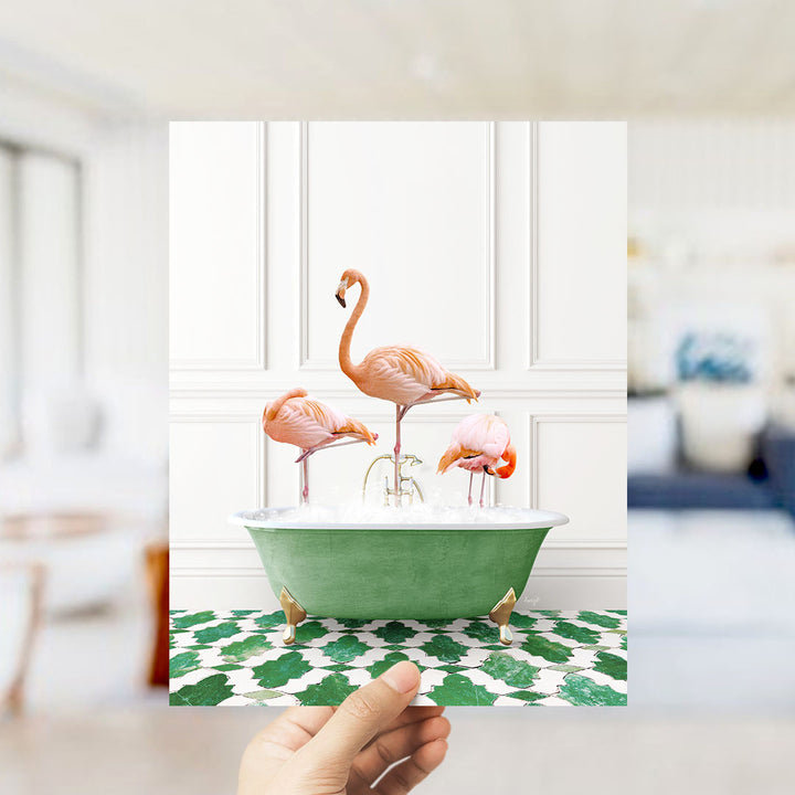 A hand holds up a framed photograph of three flamingos sitting in a green bathtub on a green and white patterned floor.