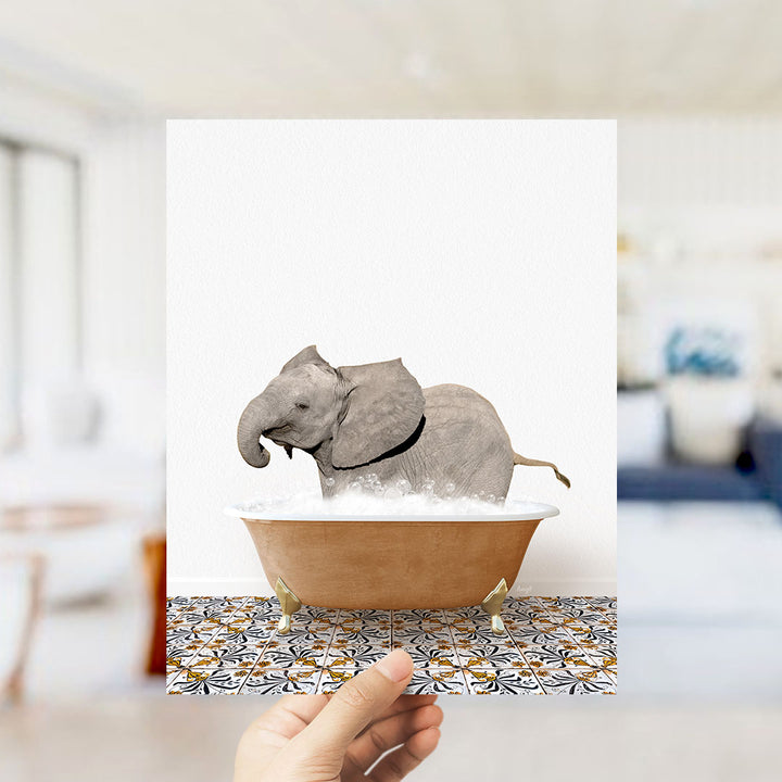A hand holds up a framed image of a baby elephant sitting in a bathtub filled with water, surrounded by a patterned tile floor.