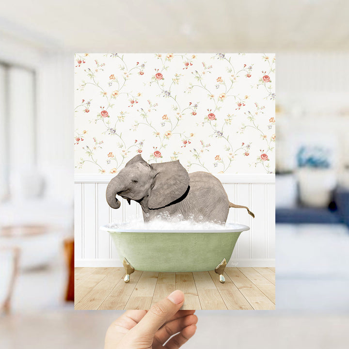 A hand holds up a framed image depicting a gray elephant sitting in a green bathtub filled with bubbles, surrounded by a floral wallpaper.