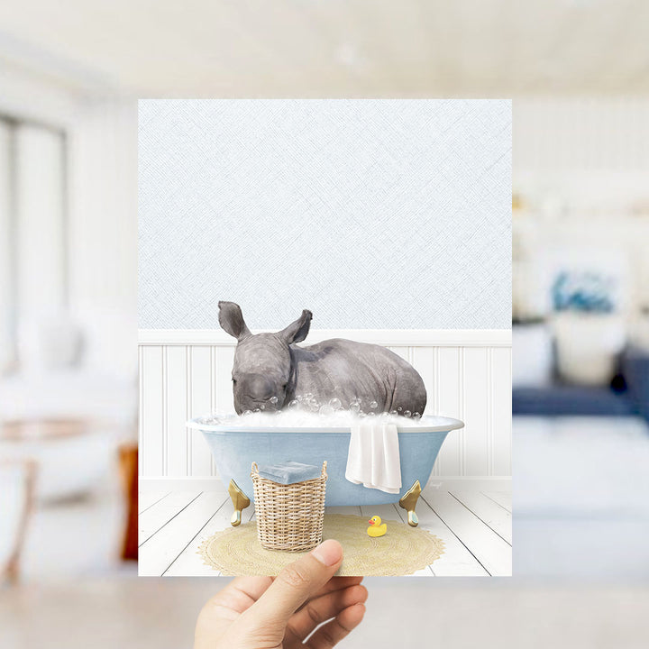 A rhinoceros is sitting in a bathtub filled with bubbles, surrounded by a yellow rubber duck and a wicker basket.