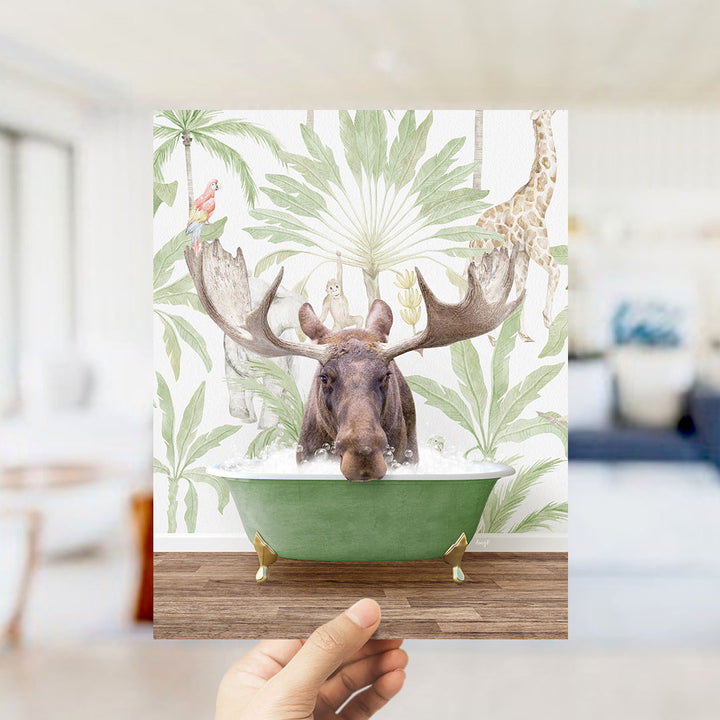 A hand holds up a collage of an image featuring a moose in a bathtub surrounded by tropical plants and animals, including a giraffe and a parrot.