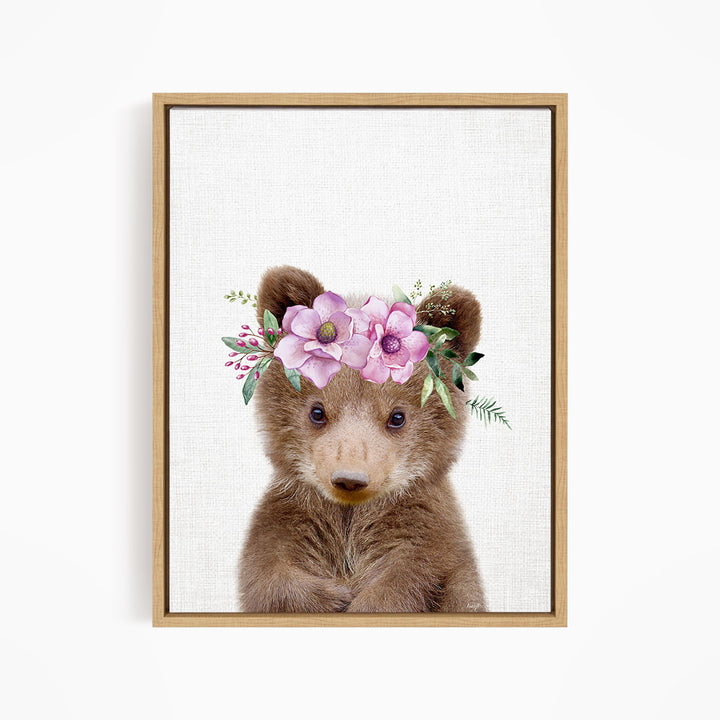 A cute brown bear wearing a floral crown on its head, with pink flowers and greenery surrounding it.