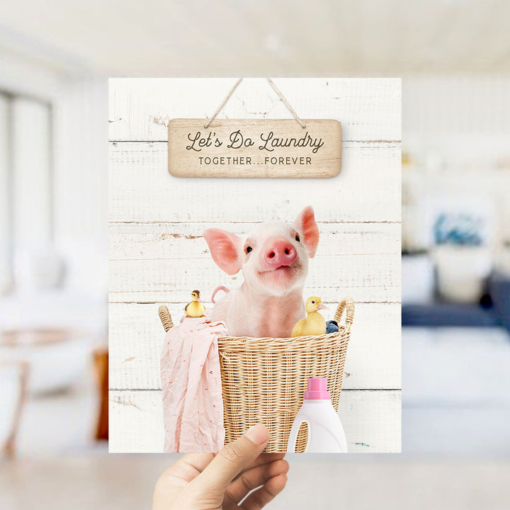 A hand holding a card with a pig in a basket and various household items, such as a bottle and a bottle cap, along with a sign that says "Let's Do Laundry Together Forever".