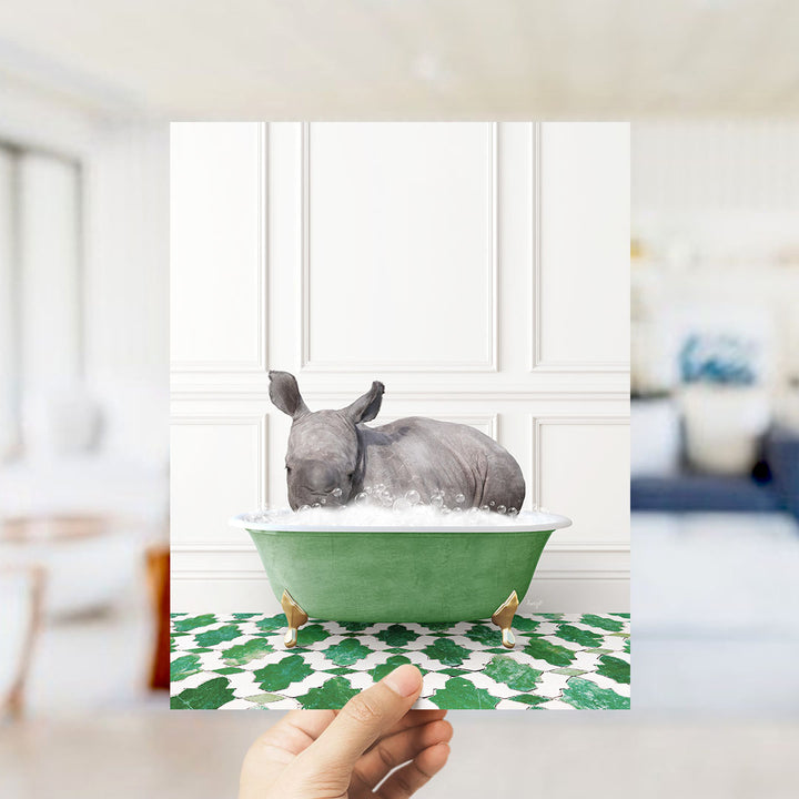 A hand holds up a framed image of a rhinoceros sitting in a green bathtub filled with bubbles, surrounded by a green and white patterned floor.