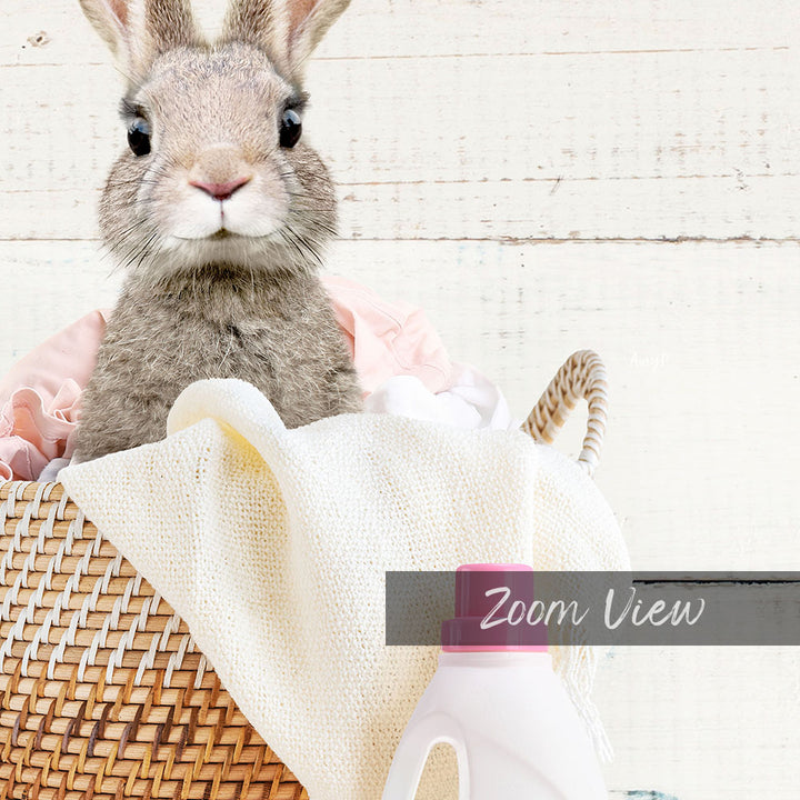 A cute gray rabbit peeking out from a woven basket filled with laundry, including a white towel and a white bottle.