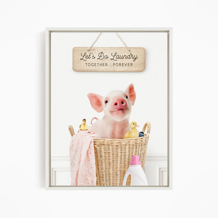 A cute pig sitting in a wicker basket filled with various items, including a pink blanket, rubber duckies, and a sign that says "Let's Do Laundry Together Forever".