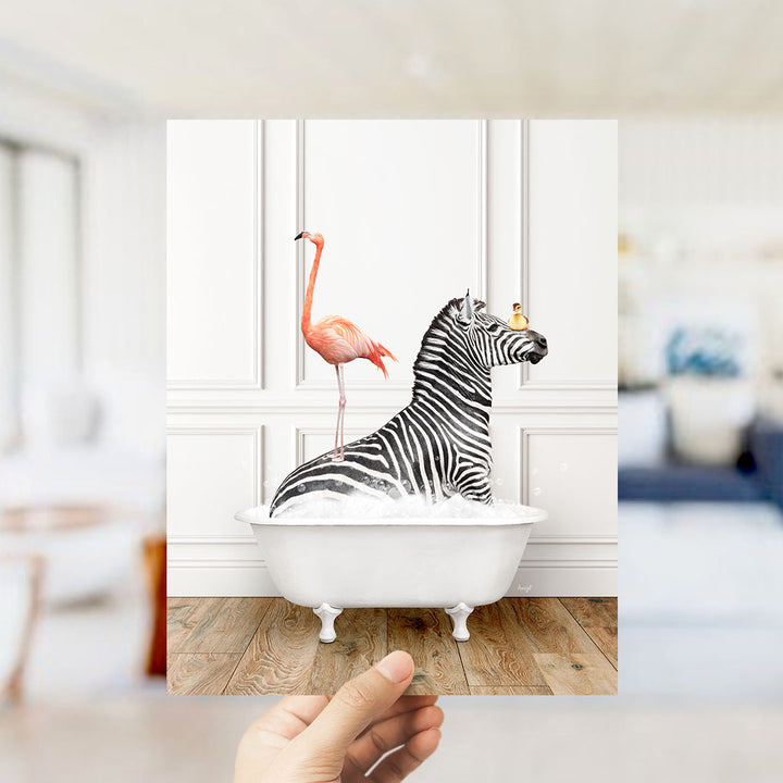 A hand holds up a framed image depicting a zebra sitting in a bathtub, with a pink flamingo standing on its back.