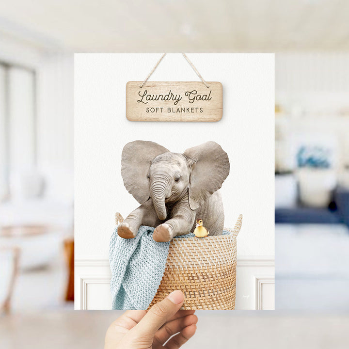 A hand is holding up a framed image of a baby elephant sitting in a woven basket, with a sign above it that says "Laundry Goal Soft Blankets".