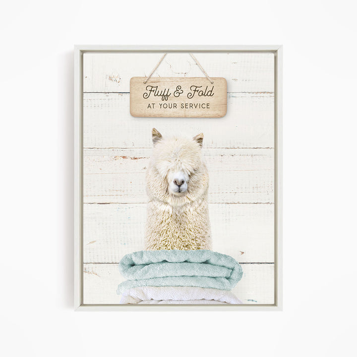 A fluffy alpaca sitting on a stack of blue towels, with a wooden sign above that says "Fuffy & Fold at your service".