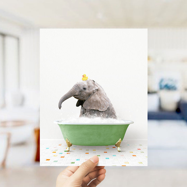 A hand holds up a framed image of a baby elephant sitting in a green bathtub, with a yellow rubber duck floating on its head.