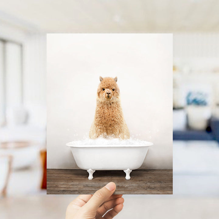 A fluffy brown alpaca sitting in a white bathtub filled with bubbles, with a blurred background suggesting a bathroom setting.