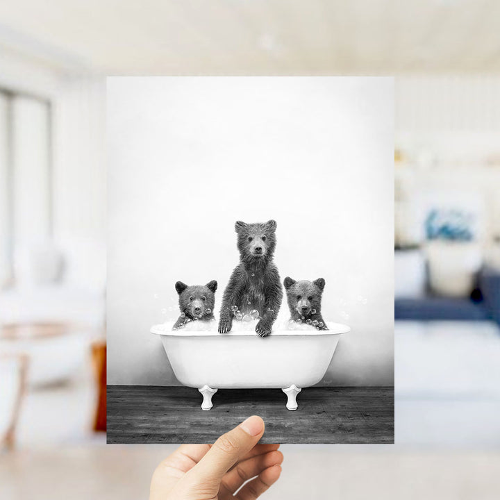 A hand holds up a framed photograph of three bears sitting in a bathtub, with the bears appearing to be in a bathtub.