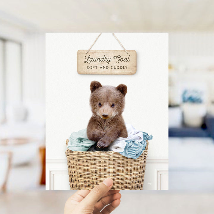 A hand holding a sign that says "Laundry Goal Soft and Cuddly" above a wicker basket containing a small brown bear.