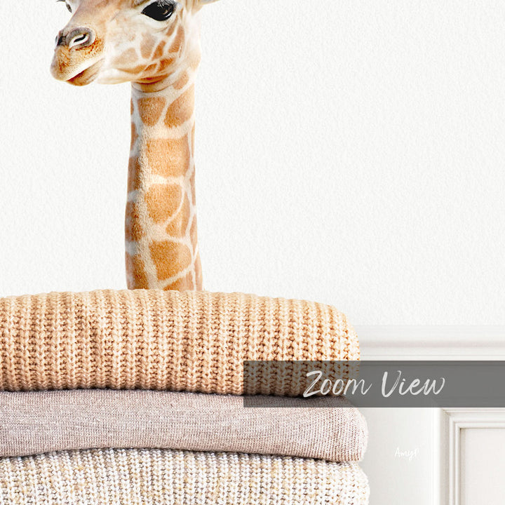 A giraffe peeking out from behind a stack of beige and brown blankets, with the text "Zoom View" overlaid on the image.