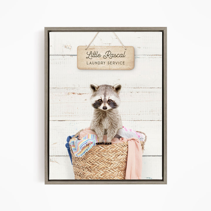 A raccoon sitting in a woven laundry basket, with a sign above that reads "Little Racal Laundry Service".