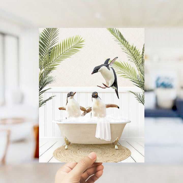 A hand holds up a framed image depicting two penguins sitting in a bathtub, surrounded by palm leaves, creating a whimsical and playful scene.