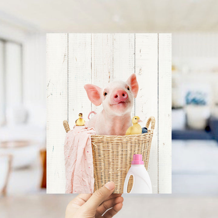 A hand holds up a framed photograph of a pig sitting in a wicker basket filled with rubber ducks.