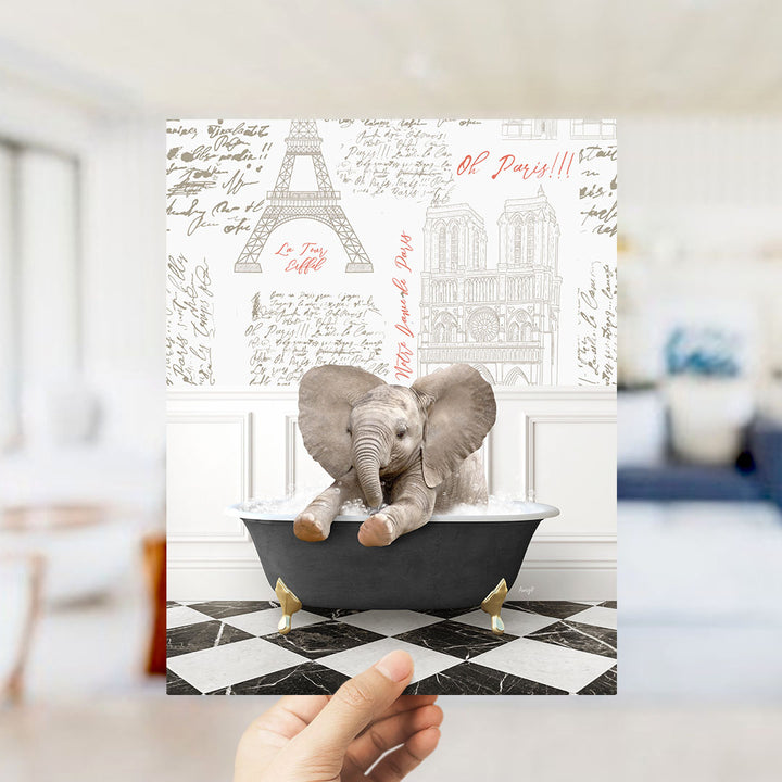 A hand is holding up a printed image featuring a small elephant sitting in a bathtub, with the Eiffel Tower and other Parisian landmarks visible in the background.