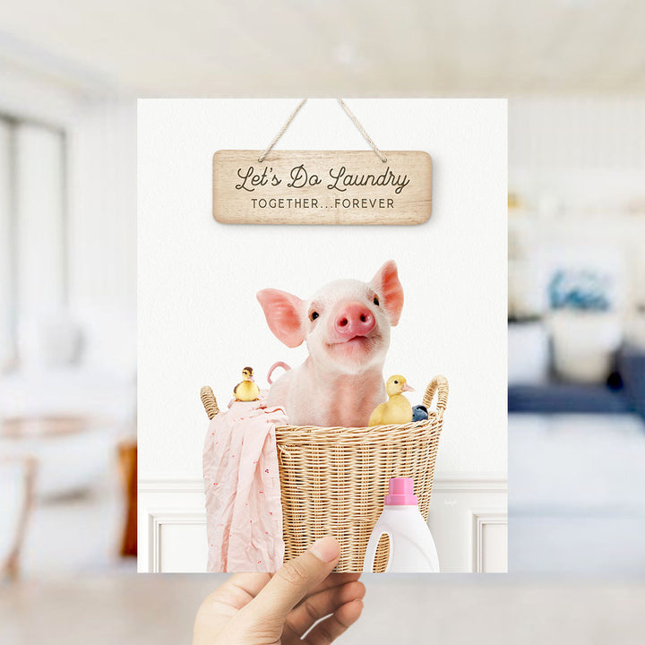 A hand holding a sign that says "Let's Do Laundry Together Forever" above a basket containing a pig and several rubber ducks.
