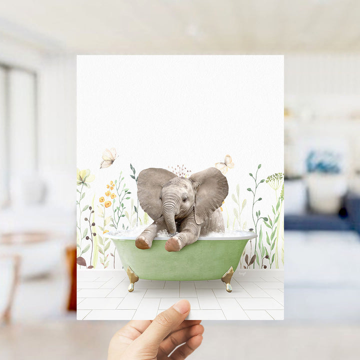 A hand holds up a framed image depicting a baby elephant sitting in a green bathtub, surrounded by flowers and butterflies.