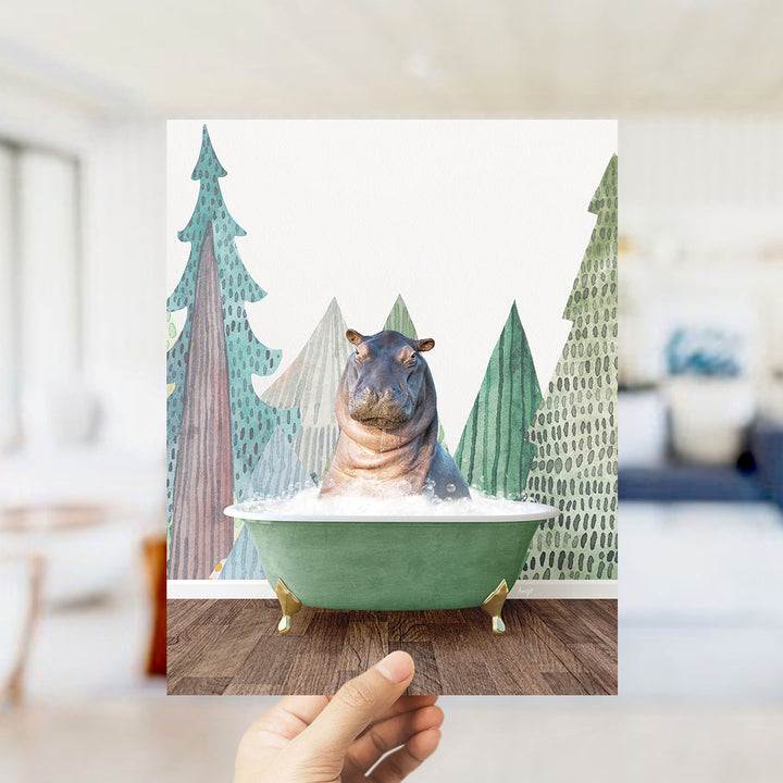 A hand holds up a framed image depicting a hippopotamus sitting in a green bathtub, surrounded by a forest-like background with trees and a snowy landscape.