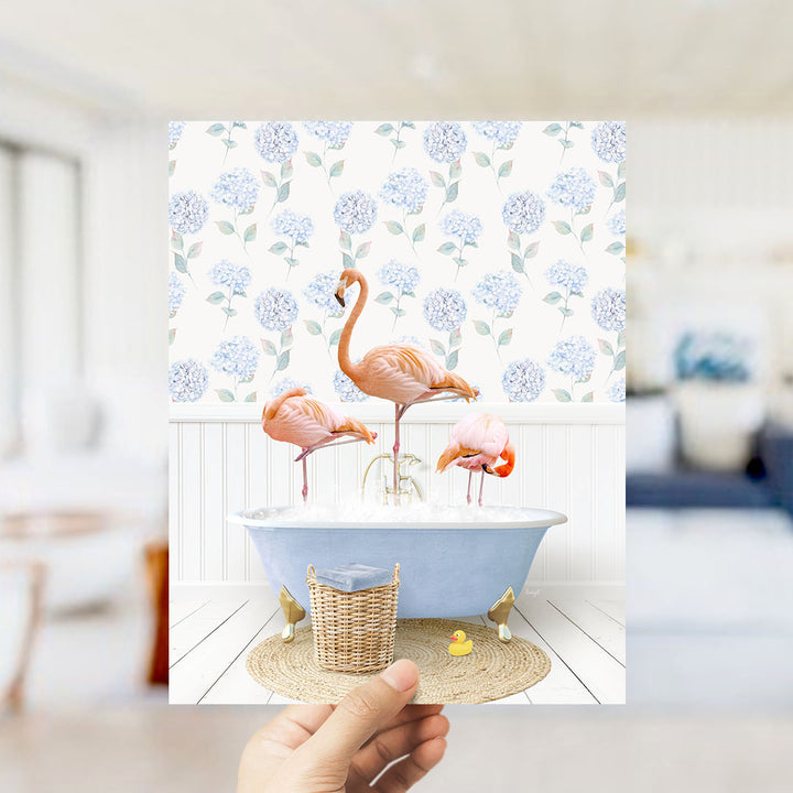 A hand holds up a framed image depicting three flamingos in a blue bathtub, surrounded by a floral patterned wall.