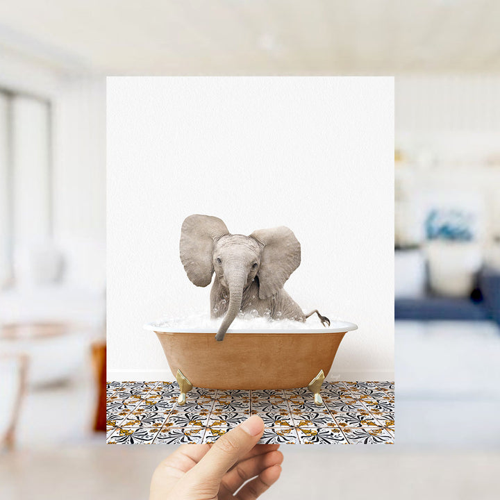 A hand holds up a framed image of a baby elephant sitting in a bathtub filled with water, surrounded by a patterned tile floor.