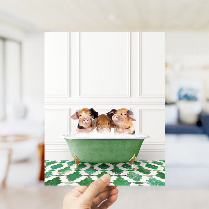 A hand holds up a framed image of three adorable pigs sitting in a green bathtub filled with bubbles, surrounded by a patterned floor.