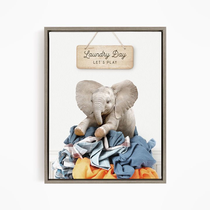 A framed image of a stuffed elephant sitting atop a pile of laundry, with a wooden sign above that reads "Laundry Day Let's Play".