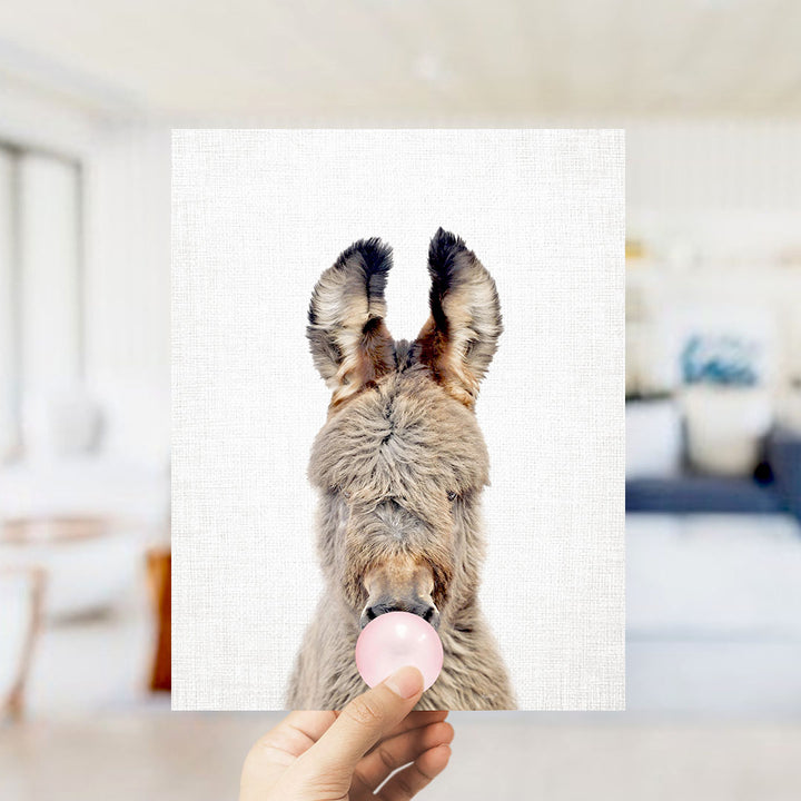 A hand holding a picture of a donkey blowing a bubble with a pink bubble gum bubble.