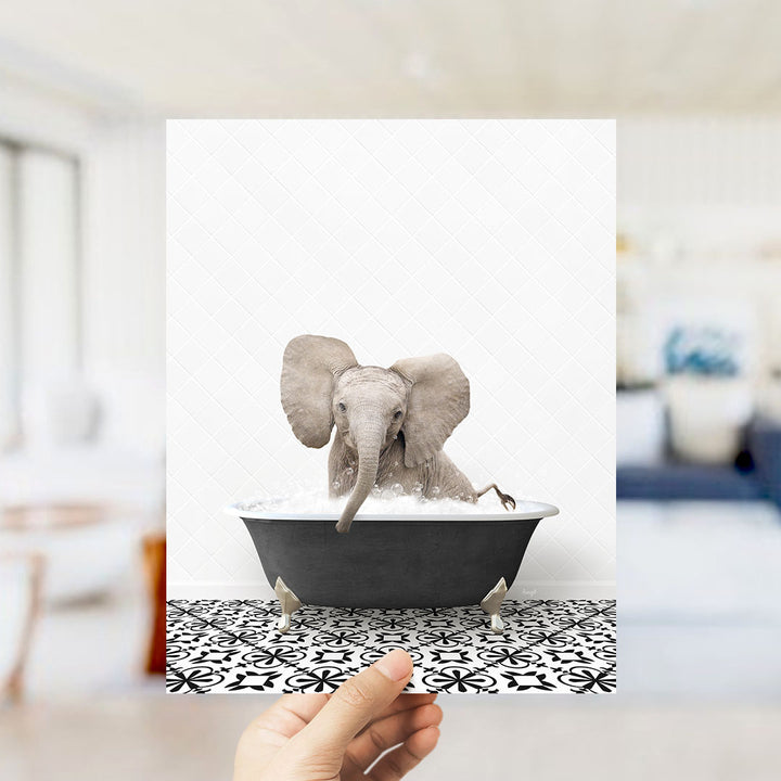 A hand holds up a framed image of a baby elephant sitting in a bathtub filled with water, surrounded by a black and white patterned floor.