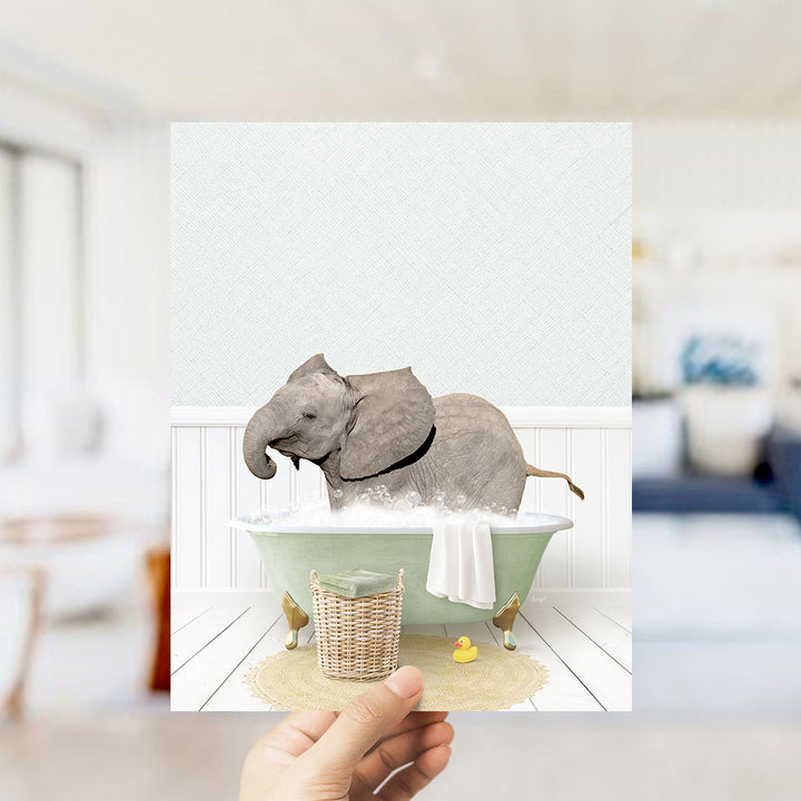 A hand holds up a framed image depicting a baby elephant enjoying a bath in a green bathtub, surrounded by a yellow rubber duck and a white towel.