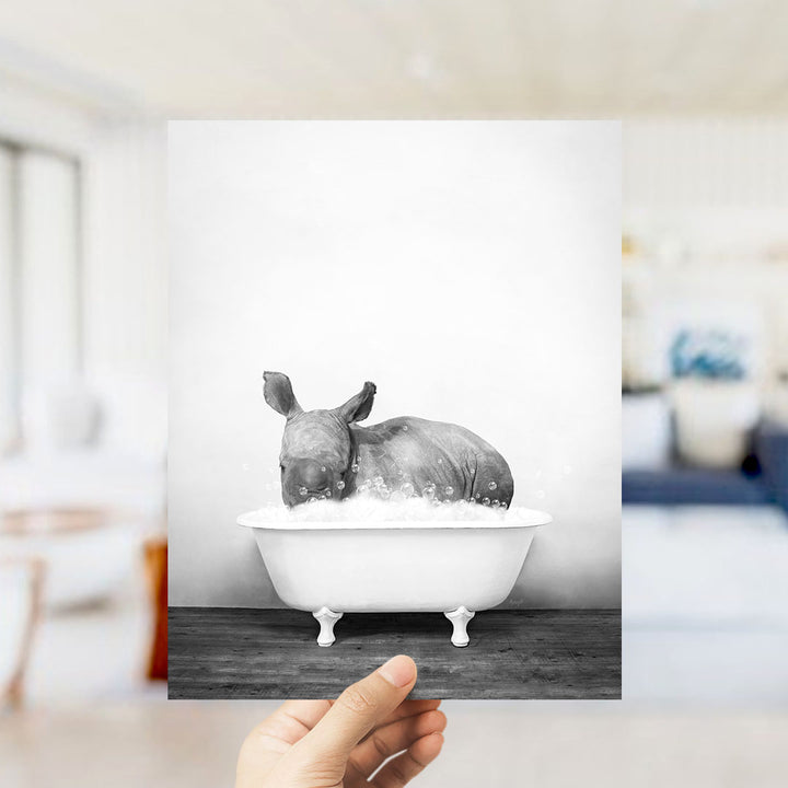 A hand holds up a black and white photograph of a rhinoceros sitting in a bathtub filled with water, creating a whimsical and playful scene.