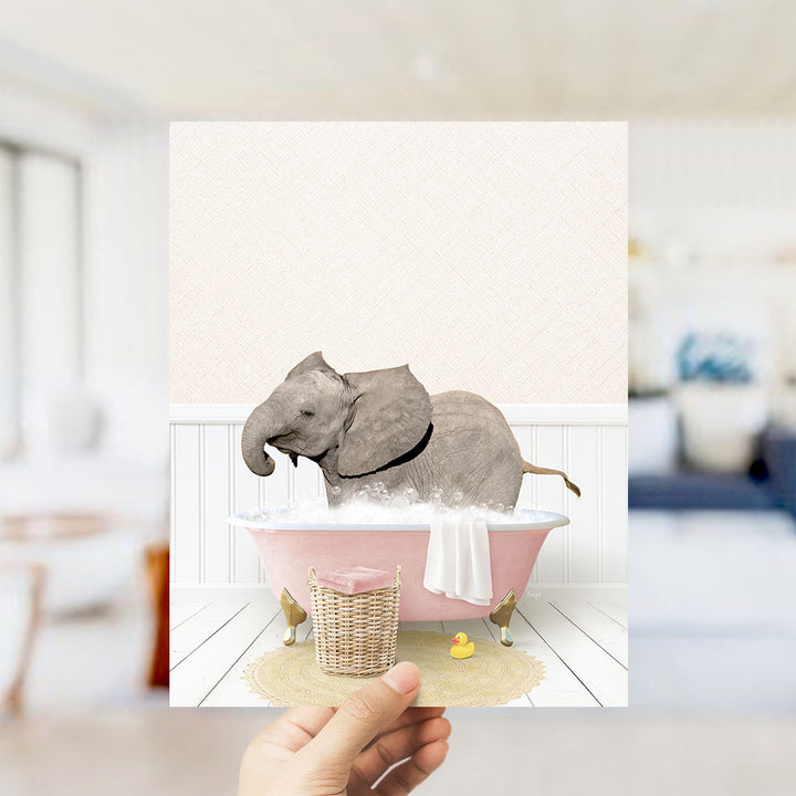 A hand holds up a framed image depicting a baby elephant sitting in a pink bathtub filled with bubbles, surrounded by a yellow rubber duck and a white towel.