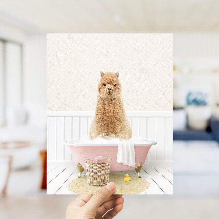 A fluffy brown alpaca sitting in a pink bathtub filled with bubbles, surrounded by a yellow rubber duck and a white towel.