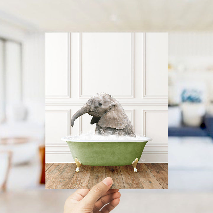 A hand holds up a framed photograph of a baby elephant sitting in a green bathtub filled with water, creating a whimsical and playful scene.