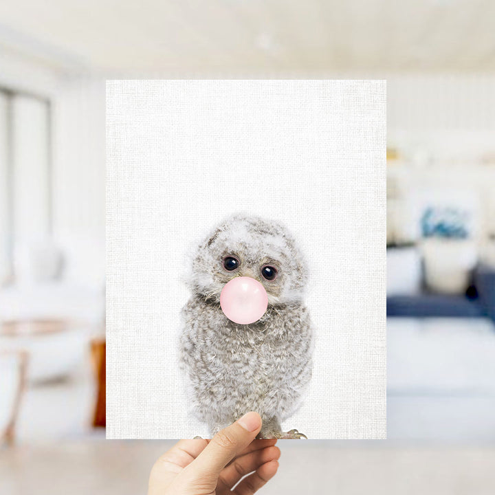 A hand is holding up a white square with a small gray owl blowing a bubble with a pink bubble gum bubble.