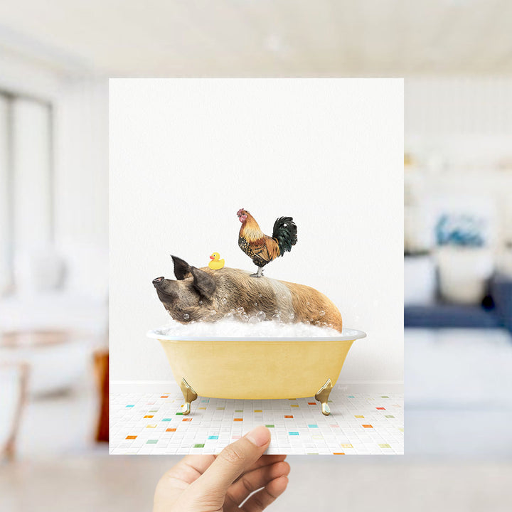 A hand holds up a framed image depicting a pig sitting in a bathtub, with a rooster perched on its back.