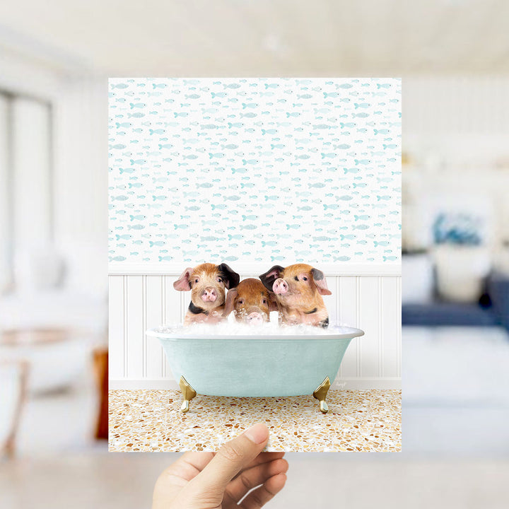 A hand holds up a framed image of three adorable pigs sitting in a bathtub filled with bubbles, surrounded by a pattern of small fish.