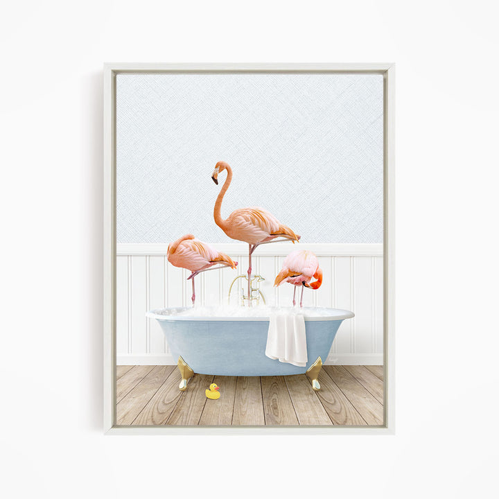 A framed image depicting three pink flamingos standing on the edge of a blue bathtub, with a white towel and rubber duckies on the floor in front of the tub.
