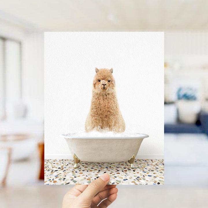 A fluffy brown alpaca sitting in a white bathtub filled with bubbles, with a hand holding the photo up to the camera.