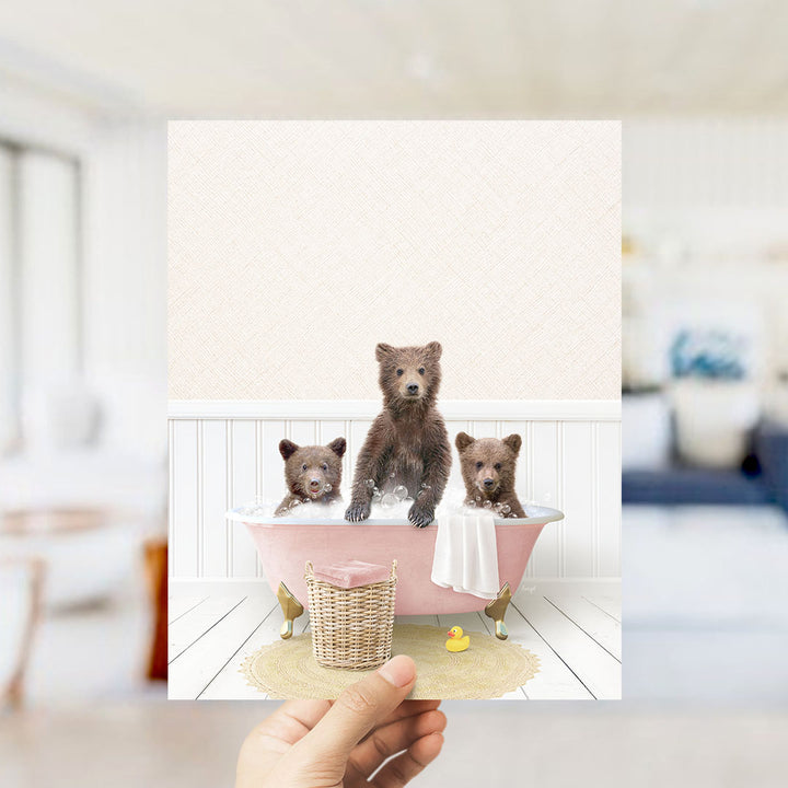 A hand holds up a framed image depicting three adorable brown bears sitting in a pink bathtub, surrounded by a yellow rubber duck and a white towel.