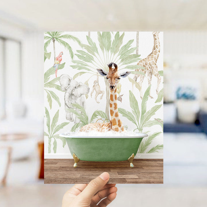 A hand holds up a framed print featuring a giraffe sitting in a bathtub surrounded by tropical plants and animals, including a parrot, elephant, and monkey.