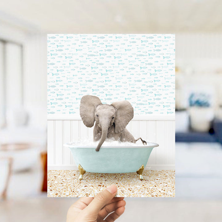 A hand holds up a framed image of a baby elephant sitting in a bathtub filled with bubbles, surrounded by a pattern of small fish.