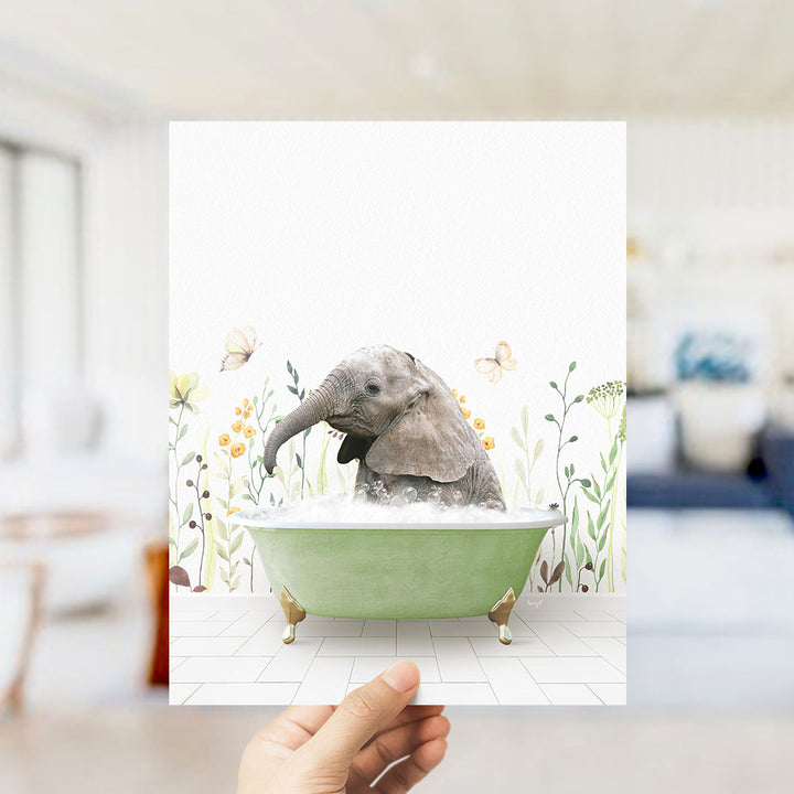A hand holds up a framed image depicting a baby elephant sitting in a green bathtub filled with bubbles, surrounded by flowers and butterflies.