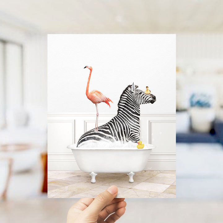 A hand holds up a framed image depicting a zebra sitting in a bathtub with a pink flamingo standing on its back.